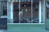 Photograph of a glass picture-window of a storefront, in which the glass reveals a view of demolition happening inside the storefront, while it also reflects the image of the exterior of a building across the street. To the left and right side of this large picture-window are smaller windows. The window to the left reflects or reveals nothing, while the window to the right reflects some light.