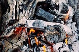 A bundle of wood charcoal, covered in white ash and orange hued flames running throughout