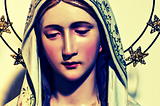 A porcelain Madonna statue with rosy red cheeks, surrounded by a halo of stars. Dominant colors of tawny yellow, green, pink, and blue shadow.