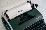 A black typewriter with a sheet of paper in it that has the word, “News” typed on it.