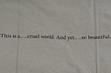 “This is a… …cruel world. And yet… …so beautiful…” printed on the back of a grey cotton tee shirt.