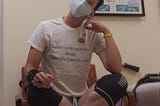 A portrait of me sitting cross-legged in a chair in a clinical exam room. I’m white-appearing with short dark hair. I’m wearing glasses, a light blue face mask over an N95, a t-shirt that says, “supercalifragilistic axial spondyloarthritis,” shorts and compression knee sleeves.