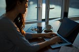 A woman sat at a laptop with a cup of coffee and pair of glasses nearby