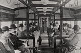 The interior of a train carriage filled with passengers, most of whom are men wearing suits and bowler hats. Despite the scene clearly being old-fashioned it is also reminscient of a modern train.