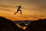 Woman jumping high between two hills, silhouetted against a sunset.
