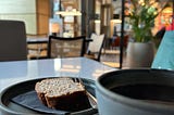 In a cafe. A close up of a dark plate with a slice of banana loaf, and the edge of a coffee cup.