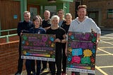 Seven people, men and women, standing together in front of a building at MDP Headquarters and those standing at the front are holding large posters advertising the Menopause Cafe