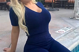 A woman with a light complexion and long blond hair. She’s wearing a blue dress, sitting on the side of a pool. She’s looking at the camera. Behind her is some patio furniture.