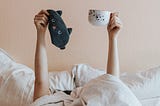 A person lying in bed has their hand raised holding an eye mask and a mug