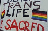 A white banner, with the red yellow and black Aboriginal flag on the left and the rainbow flag on the right with black and brown stripes, saying “black trans lives are sacred.”