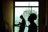 Two construction workers in shadow before a window. Improvement is ongoing.