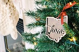 A Christmas tree in front of a fireplace with a wooden ornament in the shape of a state with the word “home,” written on it