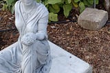 Off-white stone statue of Greek-style goddess with plant in hand in a tended garden.