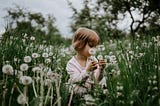 A photo of a girl sitting in a field of dendelions during daytime.