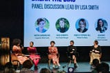 A diverse group of women sit on a stage in front of a screen. They’re participating in a panel discussion about bias on International Women’s Day 2022. Photo credit: Krysta Guille.