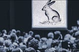 A drawing of a rabbit on a large screen in front of a crowd of people.