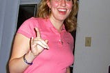 Photo of a woman with blonde hair and blue eyes wearing a pink polo shirt and making bull horns with her right hand while smiling