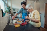 A young person with black short hair is standing in a kitchen with an elderly person with grey hair. The young person is wearing an apron, and helping serve fruit into bowls.