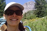 A picture of the author wearing a backpack with a field of green and a mountain in the background.