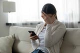 Woman sitting on the couch with a phone in her hand and her hand over her face.