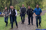 A picture of a group composed of Ithaca Common Council member Cynthia Brock, U.S. Congressperson Marc Molinaro, Tompkins County Legislator Mike Sigler, and Ithaca Police Department officers Thomas Condzella & John Joly walking down a wooded path. Some camping gear is seen at the edges of the image. Source: Local Electeds, Candidates Tour Homeless Encampment sites In Ithaca
