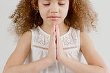 Photo of a young girl with curly hair, eyes closed and hands in “namaste” pose