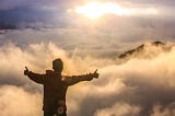 Silhouette of a young man seen from behind with arms spread, giving thumbs up as he floats in the sky amidst the clouds and the sun.