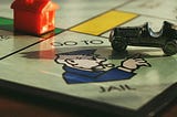 A close-up photo of a Monopoly game board, with a tiny car gamepiece resting on the “Go to Jail” space. A house gamepiece rests on the green property square next to it.