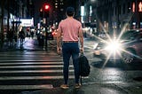 A back-view of a fit man (182 cm, 80 kg) wearing a pink t-shirt, navy blue mom-style pants, brown shoes, and a navy cap, standing calmly at a city crosswalk at night. In front of him, a speeding car flashes its bright high-beam headlights, defying the red traffic light, casting a sharp lens flare across the dark, wet street architecture.