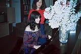 Two twenty-something sisters at a console piano, one playing and the other standing behind, singing, in a living room.
