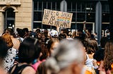 Part of the crowd at a Pride Parade. In the center is a person holding up a sign, which says “Life is short be as queer as you want” in all caps. The word queer is in the colors of the rainbow. The person standing next to them is wearing a white baseball cap with rainbow hearts on it.