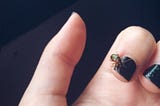 Parte de uma mão semicerrada, de que se veem duas unhas pintadas em preto e, sobre uma, um pequeno besouro dourado-esverdeado