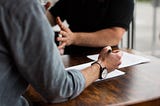 Two men talking at a conference table, while one man captures notes