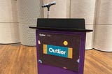 The podium at Outlier 2024 with a microphone and a sign that says “Outlier” on the front