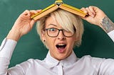 Excited young blonde female teacher wearing glasses in classroom standing in front of chalkboard holding book on head looking at camera