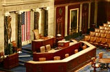 An interior view of the chamber of the US House of Representatives in the US Congress with an American flag displayed prominently at the front. The room features ornate woodwork, leather chairs arranged in rows, and a large desk with multiple chairs behind it. There is a portrait on the wall to the right and marble columns framing the flag. The carpet has a patterned design.