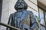 A photo of the Frederick Douglass Statue at the New York Historical Society, standing tall, proud and holding a book in his left hand close to his body.