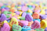 Photo of bright colored heart shaped candies, known as “conversation hearts” and usually sold around Valentine’s Day. They say things like “first kiss”, “true love” and “say yes”