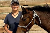 Whisper, a brown horse, and Nancy, a woman in jeans and a baseball cap, stand outside together in the sun.