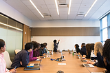 A group of diverse people sitting at a conference table. A woman stands at the front of the room point to a white board.