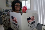 A picture of me, Aria, sitting at a table with a voting booth that says “Vote from home booth 2020.”