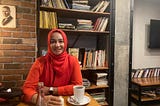 The writer, Anisa Khalifa, dressed in a brightly colored hijab enjoying a beverage near a bookshelf at a coffee shop while on vacation in Turkey.