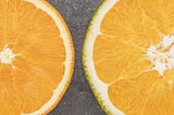 Five cross sections of citrus fruits arranged in ascending order of diameter on a textured grey background. From left to right, they appear to be a lime, a lemon, and orange, a larger orange citrus fruit, and a red grapefruit.