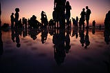 Group of people gathered at a beach at dusk, their silhouettes reflecting in the water.