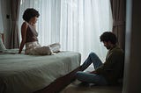 A young couple sits apart in a bedroom, avoiding each other after an argument.
