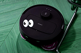 Photo of a round black robot vacuum cleaner on a floor. A googly eyes emoji has been added to the image of the vacuum cleaner.
