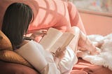 girl with dark hair wearing white shirt reading a book and sitting on pink sofa