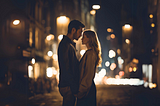 A fair-haired woman and a dark-haired man stand in a dimly lit street, gazing into one-another’s eyes.