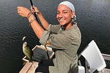 Young person of color smiling, sitting on a dock holding a fishing line with a fish she caught on the hook