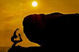 A silhouette of a young adult in an extreme yoga pose on a cliff near a giant rock with the sun in the background beginning to set in a bronze-colored sky.